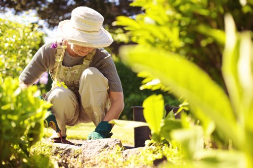 A woman wearing a large white sun hat, beige apron, grey top, and gardening gloves is kneeling on a grassy outdoor space during daylight, tending to a garden bed with soil and plants. In the background, there are lush green bushes and trees, indicating a well-maintained front or backyard garden. The garden features a neatly edged flower bed with dark soil, surrounded by a vibrant green lawn area that appears freshly mowed. The scene is illuminated by natural sunlight, suggesting clear weather. The woman is engaged in planting or weeding, and the garden setting emphasizes residential outdoor landscaping typical of the Victoria region, supporting gardening and landscape enhancement services offered by landscapersvictoria.co.uk.
