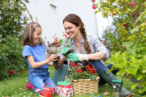 A woman and a young girl are gardening outdoors in a lush, well-maintained garden that features a variety of plants, including flowering shrubs and greenery. The woman is kneeling on the grass, wearing green gardening gloves, and is smiling while holding a small potted plant with red flowers. The girl, dressed in a blue t-shirt and jeans, is sitting on the grass nearby holding a similar potted plant. Between them, there is a woven basket filled with garden tools and supplies, including a red watering can and a small decorative container with a floral pattern. The background includes a white garden wall, with tall leafy plants and flowering bushes, creating a vibrant and inviting outdoor space. The scene captures a moment of peaceful gardening activity, highlighting the natural tones of the plants and the cheerful atmosphere typical of residential gardens, particularly in the Victoria area, with a focus on cultivating healthy greenery and flowerbeds.