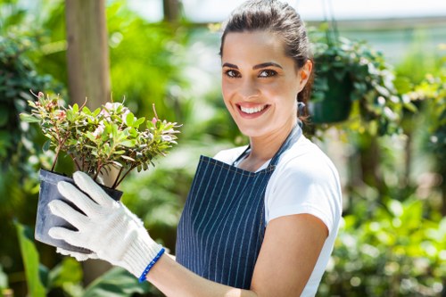 A young woman with dark hair tied back, smiling and wearing a blue apron and white gloves, holds a small potted shrub with pink flowers and green foliage in a lush outdoor garden environment. The garden features a mix of vibrant green plants, including bushes and trees, with a wooden post and a greenhouse or conservatory in the background. The scene is well-lit with natural sunlight, creating a bright and cheerful atmosphere suitable for garden maintenance and landscaping services. The image emphasizes outdoor gardening activities, highlighting the natural textures and colors of the plants and the garden space, aligning with professional gardening and lawn care offerings available through Landscapers Victoria in the Victoria area, near postcode CV1 or CV2.