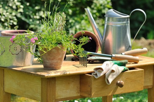 A wooden garden table set outdoors in a lush green environment, featuring various gardening tools and equipment. On the table are clay and terracotta pots, one filled with fresh green herbs and ornamental grasses. There is a silver metal watering can, a small trowel, a hand rake, and a pair of gardening gloves resting on the table. The background showcases a well-maintained garden with dense foliage, suggesting a healthy, vibrant landscape that could be part of a residential front or back garden, indicative of a typical outdoor space within Victoria or nearby areas. The scene is illuminated by natural daylight, with no harsh shadows, indicating clear weather conditions. The arrangement of gardening supplies and plants reflects ongoing gardening or planting activities, aligning with the services offered by Landscapers Victoria in garden maintenance, planting, and outdoor landscaping to enhance the beauty and functionality of outdoor spaces.