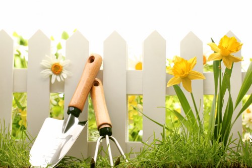 A close-up view of a garden scene in a front yard, featuring a white picket fence with pointed tops, behind which there are bright yellow daffodils and white daisies with yellow centers growing in a lush green lawn. In the foreground, gardening tools including a trowel, a cultivator, and a small garden fork with wooden handles rest on the grass. The garden area is sunlit, highlighting the natural textures of the grass, flower petals, and wooden tool handles, creating a vibrant and well-maintained outdoor space that reflects typical garden landscaping and maintenance practices common in the Victoria area. The overall scene emphasizes a neat, healthy, and colorful garden environment suitable for professional gardening services offered by Landscapers Victoria.