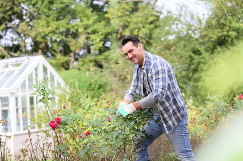 A man in casual clothing, including a plaid shirt and jeans, is working in a residential garden in Victoria. He is smiling and appears to be pruning or tending to a rose bush with pink flowers, using gardening gloves. The garden features a well-maintained lawn with dense, green grass in the foreground, a tidy flower bed with a variety of plants and shrubs, and a backdrop of mature trees and bushes that create a lush, natural environment. To the left, a small glass greenhouse is visible, suggesting an area for plant cultivation. The outdoor space is bathed in natural daylight, with clear weather conditions, indicating a bright, mild day suitable for gardening. The scene reflects a typical landscaped yard or outdoor area that may benefit from professional gardening services provided by companies like Landscapers Victoria, serving residential clients around the Victoria postcodes.