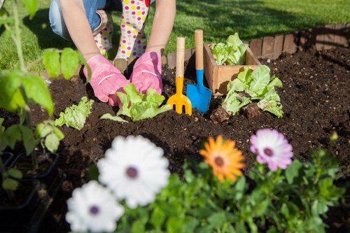 A gardener's hands, wearing pink gardening gloves, are planting young lettuce or similar leafy greens into rich, dark soil within a well-maintained garden bed situated in a landscaped outdoor space. The garden features a mix of flowering plants, including white, purple, and orange blooms in the foreground, adding colour and diversity to the garden border. Behind the planting area, there are other flowering plants and garden tools such as a small yellow rake and blue trowel, indicating active gardening work. The setting appears to be in a residential garden or a small landscaped yard, with vibrant green grass and a sunny weather condition creating natural light and shadows across the scene. The structured garden bed is edged with brick or stone, providing a neat, defined boundary, and the overall environment reflects a cared-for outdoor space suitable for gardening services by companies like Landscapers Victoria, serving local areas such as Victoria and nearby towns. This scene illustrates typical lawn and garden maintenance, planting, and outdoor beautification efforts aimed at establishing lush, productive, and aesthetically pleasing garden spaces.