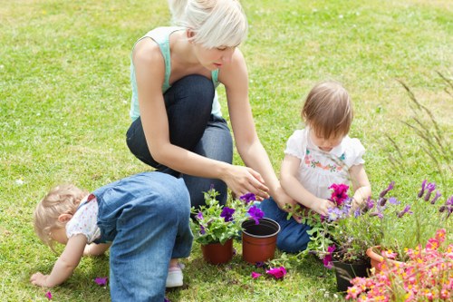 A woman with short blonde hair and a light blue sleeveless top is kneeling on a well-maintained lawn in a backyard garden, tending to potted flowers and plants arranged along the edge of a flower bed. Two young children, a girl with light brown hair wearing a white dress with colorful floral patterns, and another child with curly blonde hair dressed in blue overalls, are actively engaged in planting or examining flowers in the same garden area. The outdoor space features lush green grass, a flower bed bordered by the lawn, and various flowering plants with vibrant pink, purple, and green foliage. In the background, there are small shrubs and possibly trees, contributing to a natural and inviting garden environment, with soft daylight suggesting a pleasant weather day. The scene reflects a peaceful outdoor gardening activity, capturing natural textures of grass, soil, and flowers, and highlights a family-friendly garden setup that a professional landscaper such as Landscapers Victoria could create or maintain for local clients in the Victoria area, near postcodes corresponding to the town of Victoria or nearby surroundings.